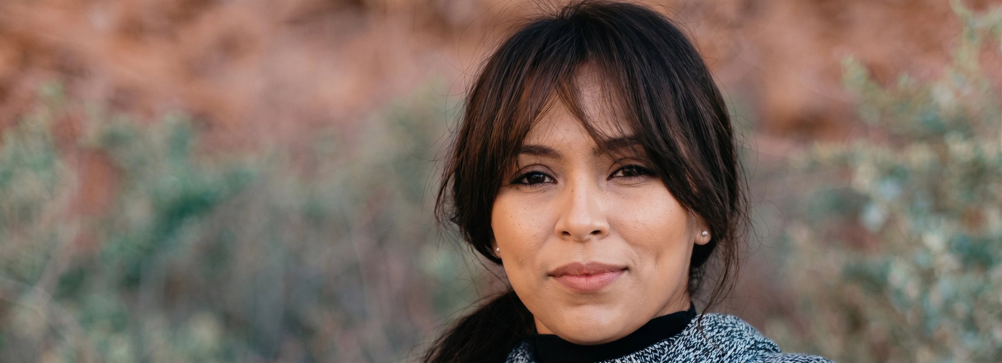 A close-up portrait of a woman with dark hair and bangs, wearing a textured gray knit top over a black collar, set against a blurred natural background of reddish rocks and greenery.