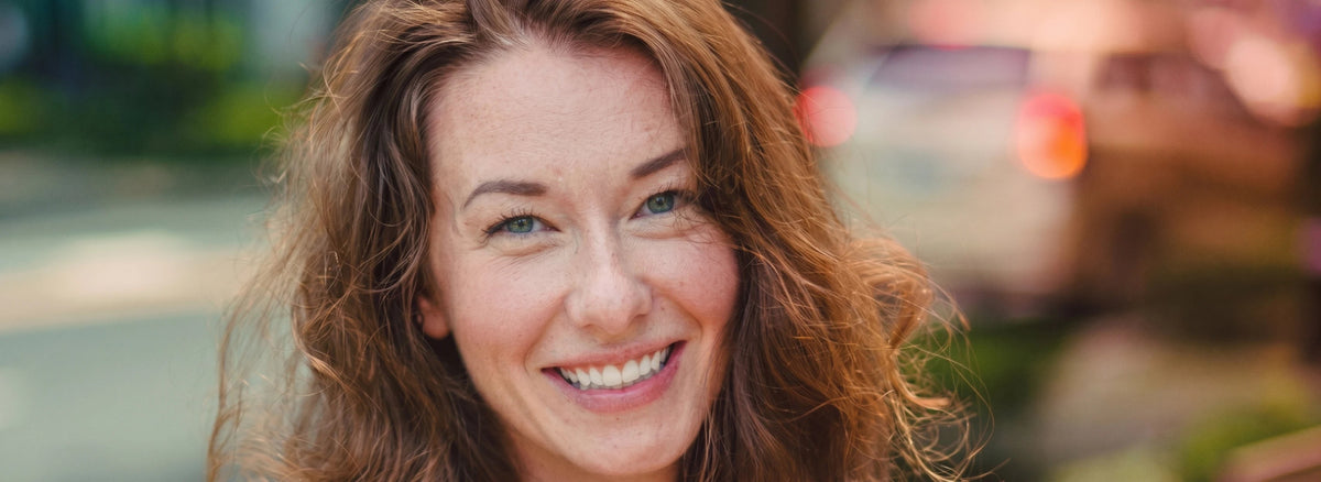 A close-up portrait of a smiling woman with wavy light brown hair, shown outdoors with a blurred background.