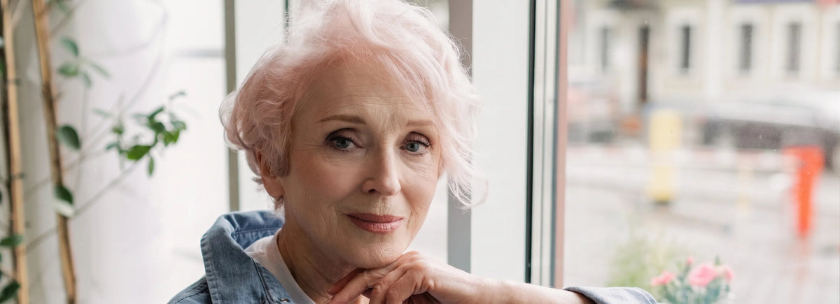 An older woman with short pink hair resting her chin on her hand, wearing a denim jacket, seated in front of a window with a blurred outdoor street scene in the background.